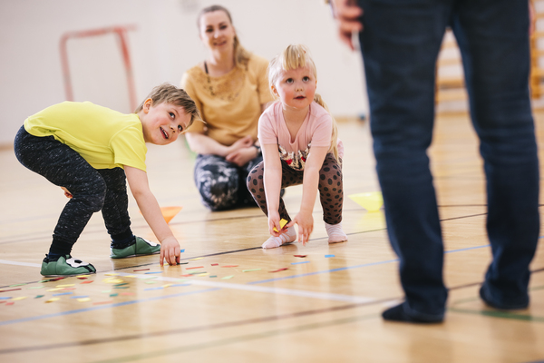 Hymyilevät lapset keräävät liikuntasalin lattialta värikkäitä paperilappuja. Taustalla aikuinen katsoo lasten leikkejä.
