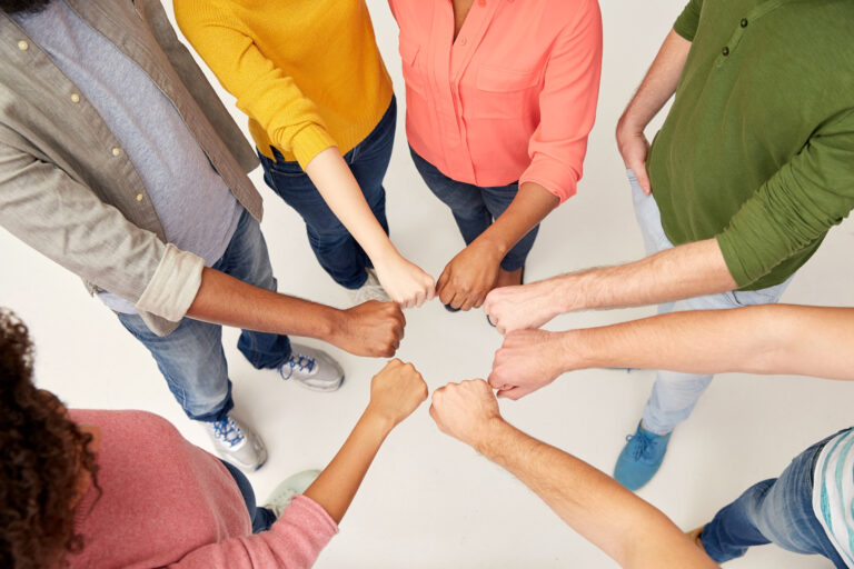 Seitsemän ihmistä seisomassa ympyrässä, kädet yhdessä keskellä yhteistyön merkiksi tekemässä ns. nyrkkitervehdyksen.