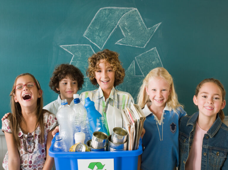 Lapsia kierrätysastian äärellä. Astiassa muovipulloja, metallitölkkejä ja sanomalehtiä.