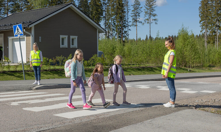 Kolme lasta kävelee suojatiellä ja kaksi aikuista valvoo